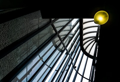 A glass roof structure featuring a series of curved, intersecting beams and transparent panels. A glowing light fixture in the upper right corner adds warmth to the otherwise cool-toned architectural elements, creating a dynamic play of lines and light.