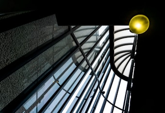 A glass roof structure featuring a series of curved, intersecting beams and transparent panels. A glowing light fixture in the upper right corner adds warmth to the otherwise cool-toned architectural elements, creating a dynamic play of lines and light.