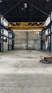 An empty industrial warehouse with high ceilings and large metal beams. The space is spacious with concrete floors and walls that show signs of wear, offering a gritty and raw aesthetic. Overhead, a big yellow crane is installed, indicating the space's potential for lifting heavy materials.