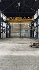 An empty industrial warehouse with high ceilings and large metal beams. The space is spacious with concrete floors and walls that show signs of wear, offering a gritty and raw aesthetic. Overhead, a big yellow crane is installed, indicating the space's potential for lifting heavy materials.