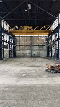 An empty industrial warehouse with high ceilings and large metal beams. The space is spacious with concrete floors and walls that show signs of wear, offering a gritty and raw aesthetic. Overhead, a big yellow crane is installed, indicating the space's potential for lifting heavy materials.
