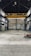 An empty industrial warehouse with high ceilings and large metal beams. The space is spacious with concrete floors and walls that show signs of wear, offering a gritty and raw aesthetic. Overhead, a big yellow crane is installed, indicating the space's potential for lifting heavy materials.