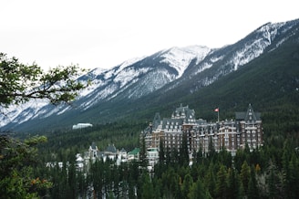 A large, historic hotel with a distinctive architectural style is nestled among dense evergreen trees with towering snow-capped mountains in the background. The hotel has ornate, castle-like features, including multiple turrets and a Canadian flag flying from one of its towers.