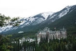 A large, historic hotel with a distinctive architectural style is nestled among dense evergreen trees with towering snow-capped mountains in the background. The hotel has ornate, castle-like features, including multiple turrets and a Canadian flag flying from one of its towers.