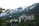 A large, historic hotel with a distinctive architectural style is nestled among dense evergreen trees with towering snow-capped mountains in the background. The hotel has ornate, castle-like features, including multiple turrets and a Canadian flag flying from one of its towers.