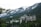 A large, historic hotel with a distinctive architectural style is nestled among dense evergreen trees with towering snow-capped mountains in the background. The hotel has ornate, castle-like features, including multiple turrets and a Canadian flag flying from one of its towers.