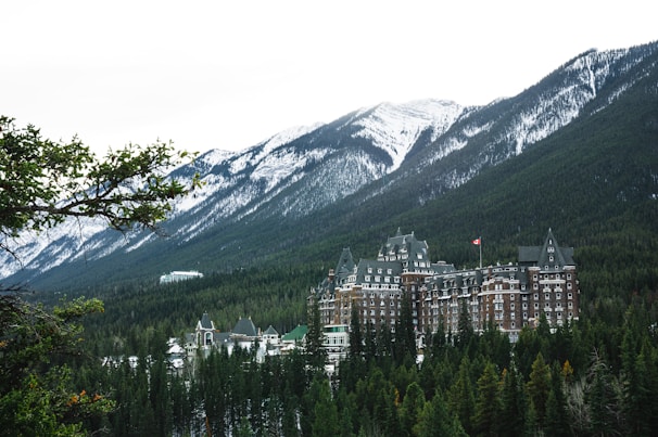 A large, historic hotel with a distinctive architectural style is nestled among dense evergreen trees with towering snow-capped mountains in the background. The hotel has ornate, castle-like features, including multiple turrets and a Canadian flag flying from one of its towers.