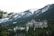 A large, historic hotel with a distinctive architectural style is nestled among dense evergreen trees with towering snow-capped mountains in the background. The hotel has ornate, castle-like features, including multiple turrets and a Canadian flag flying from one of its towers.