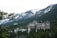 A large, historic hotel with a distinctive architectural style is nestled among dense evergreen trees with towering snow-capped mountains in the background. The hotel has ornate, castle-like features, including multiple turrets and a Canadian flag flying from one of its towers.