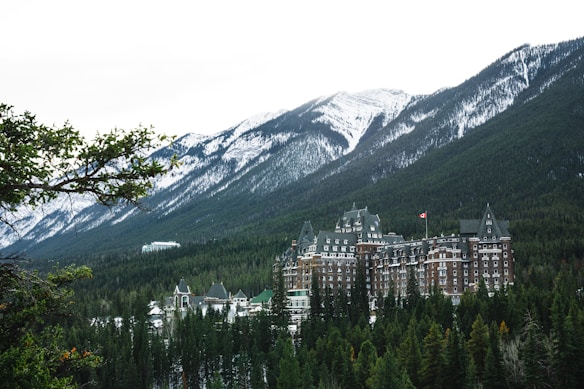 A large, historic hotel with a distinctive architectural style is nestled among dense evergreen trees with towering snow-capped mountains in the background. The hotel has ornate, castle-like features, including multiple turrets and a Canadian flag flying from one of its towers.