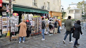People are browsing and shopping at a street market in front of a store named La Trouvaille. The shop displays various items such as postcards, t-shirts, and souvenirs. Nearby, a sign for a café named Café Montmartre is visible, with several pedestrians walking on the cobblestone street.