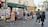 People are browsing and shopping at a street market in front of a store named La Trouvaille. The shop displays various items such as postcards, t-shirts, and souvenirs. Nearby, a sign for a café named Café Montmartre is visible, with several pedestrians walking on the cobblestone street.