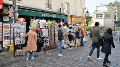 People are browsing and shopping at a street market in front of a store named La Trouvaille. The shop displays various items such as postcards, t-shirts, and souvenirs. Nearby, a sign for a caf&eacute; named Caf&eacute; Montmartre is visible, with several pedestrians walking on the cobblestone street.