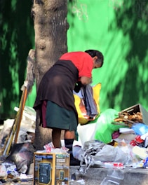 A person with dark hair, wearing a red top and dark skirt, is seen bending over a pile of trash. The setting is outdoor, next to a tree with a bright green background wall. The pile includes various items like boxes, bags, and plastic containers. A mop and walking stick are leaning against the tree.