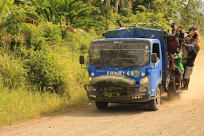 A blue truck loaded with people and cargo drives along a dirt road. The truck appears packed, with individuals sitting on the roof and hanging onto the sides, while large bundles are secured at the front. Lush green vegetation surrounds the road, indicating a rural setting in a tropical climate.