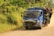 A blue truck loaded with people and cargo drives along a dirt road. The truck appears packed, with individuals sitting on the roof and hanging onto the sides, while large bundles are secured at the front. Lush green vegetation surrounds the road, indicating a rural setting in a tropical climate.