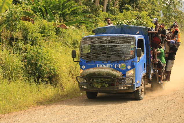 A blue truck loaded with people and cargo drives along a dirt road. The truck appears packed, with individuals sitting on the roof and hanging onto the sides, while large bundles are secured at the front. Lush green vegetation surrounds the road, indicating a rural setting in a tropical climate.