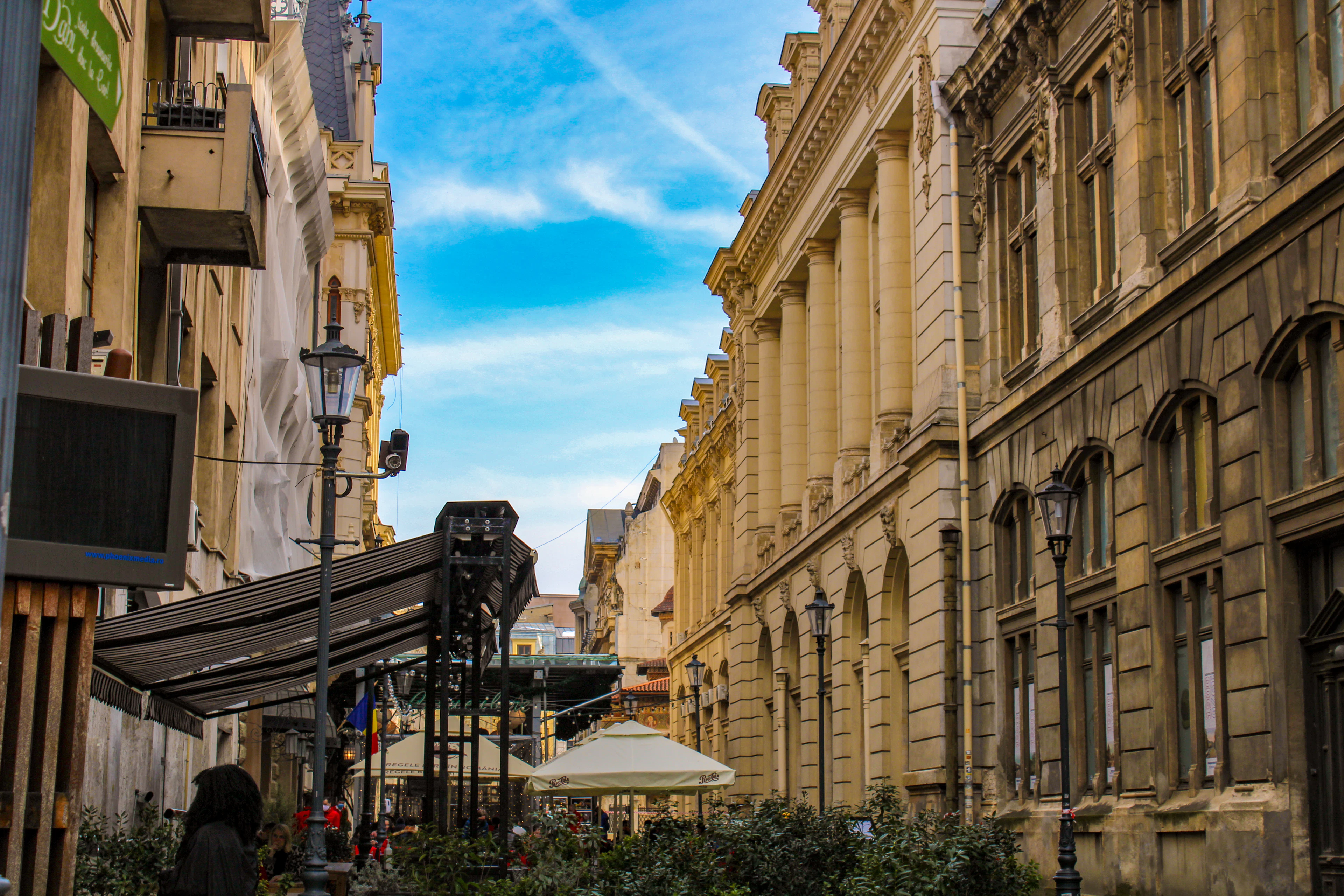 A street in Bucharest's Old Town on a bright day, a grand columned nineteenth-century stone facade running down one side and cafe awnings and parasols along the other, beneath a blue sky streaked with contrails.
