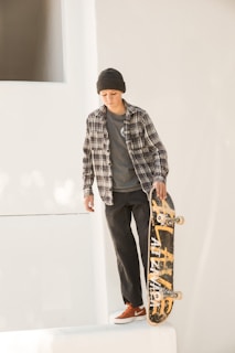 A person wearing a plaid shirt, beanie, and red sneakers is standing on a white ledge holding a skateboard vertically. The skateboard has a worn, yellow graphic design, and the background is minimalistic and white, suggesting an urban environment.