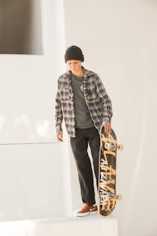 A person wearing a plaid shirt, beanie, and red sneakers is standing on a white ledge holding a skateboard vertically. The skateboard has a worn, yellow graphic design, and the background is minimalistic and white, suggesting an urban environment.