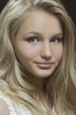 A close-up portrait of a person with long, flowing blonde hair and light eyes. The face is softly illuminated, highlighting smooth skin and a subtle smile.