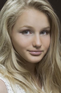 A close-up portrait of a person with long, flowing blonde hair and light eyes. The face is softly illuminated, highlighting smooth skin and a subtle smile.
