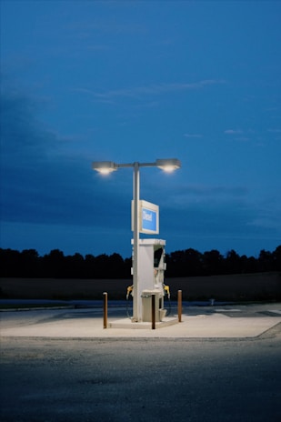 A lone diesel gas pump stands illuminated under two overhead lights against a dark blue evening sky. The area is surrounded by shadowy fields and trees in the distance, emphasizing the pump as the central focus.