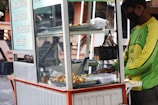 A street food vendor operates a mobile food cart, serving various fried snacks. The vendor, wearing a green shirt and a mask, is focused on preparing food. The cart displays a menu listing different flavors on the side, with containers and cooking utensils visible on the countertop.