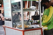 A street food vendor operates a mobile food cart, serving various fried snacks. The vendor, wearing a green shirt and a mask, is focused on preparing food. The cart displays a menu listing different flavors on the side, with containers and cooking utensils visible on the countertop.