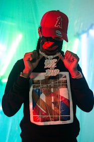 A person is wearing a red cap with an embroidered logo and a black hoodie featuring a colorful abstract design. They are also wearing a mask with a red pattern and holding up a large metallic chain necklace with the words 'HAND MADE'. The background is illuminated with green and blue lighting, creating a vibrant and edgy atmosphere.