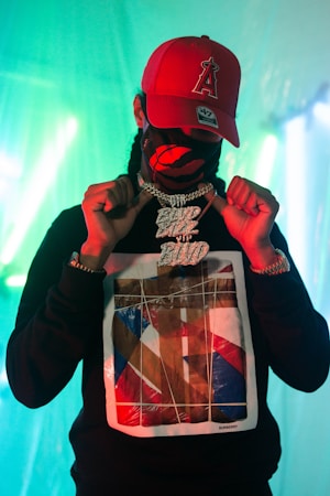 A person is wearing a red cap with an embroidered logo and a black hoodie featuring a colorful abstract design. They are also wearing a mask with a red pattern and holding up a large metallic chain necklace with the words 'HAND MADE'. The background is illuminated with green and blue lighting, creating a vibrant and edgy atmosphere.