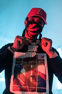 A person wearing a red cap with a symbol, a black and red patterned face mask, and a black sweatshirt with a graphic design. They are displaying a large, ornate chain necklace with shiny, bold letters. The background has a soft blue light, enhancing the overall contrast.