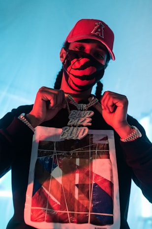 A person wearing a red cap with a symbol, a black and red patterned face mask, and a black sweatshirt with a graphic design. They are displaying a large, ornate chain necklace with shiny, bold letters. The background has a soft blue light, enhancing the overall contrast.