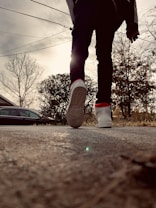 A low-angle view of a person walking on a concrete path, highlighting the sole of their white sneaker with a red detail. The person is silhouetted against a cloudy sky with visible utility lines. On the side, leafless trees and shrubs are present, with a car partially seen near the background.