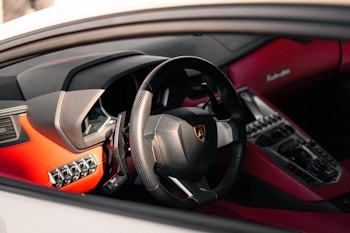 The interior of a luxury sports car featuring a sleek steering wheel with a logo, surrounded by a red and black dashboard with various controls and buttons.