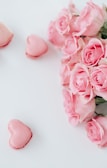 A bouquet of pink roses positioned next to two heart-shaped pink macarons on a white surface, creating a soft and romantic composition.