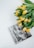 A book titled 'The Essence of Photography' with black and white forest imagery on the cover is placed next to a bouquet of yellow tulips with green leaves. The book and flowers are arranged against a clean white background, creating a minimalist aesthetic.