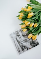 A book titled 'The Essence of Photography' with black and white forest imagery on the cover is placed next to a bouquet of yellow tulips with green leaves. The book and flowers are arranged against a clean white background, creating a minimalist aesthetic.