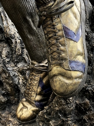 A close-up of a bronze statue depicting a pair of vintage soccer shoes with distinct laces and textured surface, standing on a rugged base.