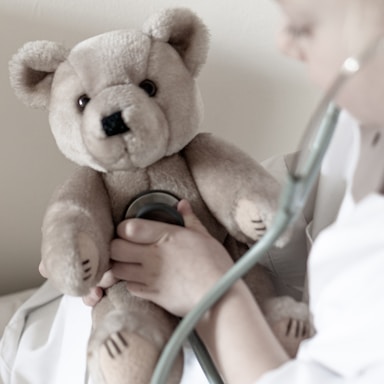 A teddy bear is being held while a person uses a stethoscope to listen to its chest. The person is wearing a white coat, suggesting they are playing a doctor or engaged in a medical role-play.
