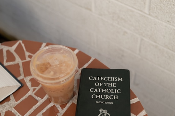 A mosaic-tiled surface holds a plastic cup of iced coffee next to a book titled 'Catechism of the Catholic Church, Second Edition'. The background features a neutral-toned wall.