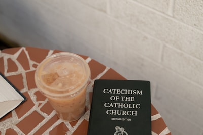 A mosaic-tiled surface holds a plastic cup of iced coffee next to a book titled 'Catechism of the Catholic Church, Second Edition'. The background features a neutral-toned wall.