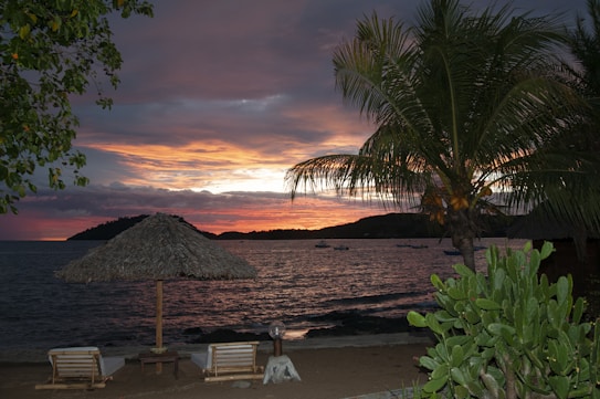 A serene beach setting during sunset with a dramatic sky, featuring hues of orange, pink, and purple blending together. The foreground contains two lounge chairs under a thatched umbrella, surrounded by lush palm trees and succulent plants. The ocean is calm, with a few small boats visible in the distance, creating a tranquil and picturesque atmosphere.