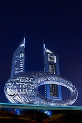 A futuristic building with an oval-shaped structure covered in Arabic calligraphy is prominently displayed. Two tall, illuminated skyscrapers stand in the background against a night sky.