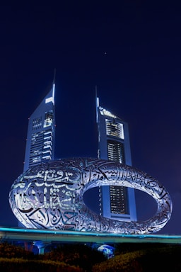 A futuristic building with an oval-shaped structure covered in Arabic calligraphy is prominently displayed. Two tall, illuminated skyscrapers stand in the background against a night sky.