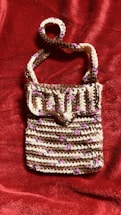 A crocheted handbag featuring a striped pattern with alternating rows of brown, white, and small pink accents. The bag has a flap closure and a single strap, displayed against a textured, deep red fabric background.