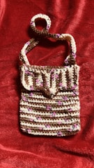 A crocheted handbag featuring a striped pattern with alternating rows of brown, white, and small pink accents. The bag has a flap closure and a single strap, displayed against a textured, deep red fabric background.