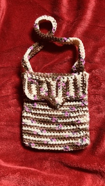 A crocheted handbag featuring a striped pattern with alternating rows of brown, white, and small pink accents. The bag has a flap closure and a single strap, displayed against a textured, deep red fabric background.