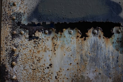A close-up view of an old, rusted metal surface with peeling paint. The corrosion has eaten through parts of the surface, creating an uneven, textured appearance with varying shades of rust, blue, and gray. The metal is bolted, and the bolts themselves are corroded.