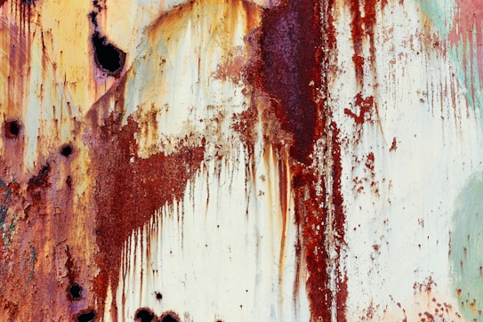 Corroded metal surface with streaks of reddish-brown rust cascading downwards, interspersed with patches of creamy off-white paint and dark spots. The texture is rough and degraded, giving an impression of natural decay over time.